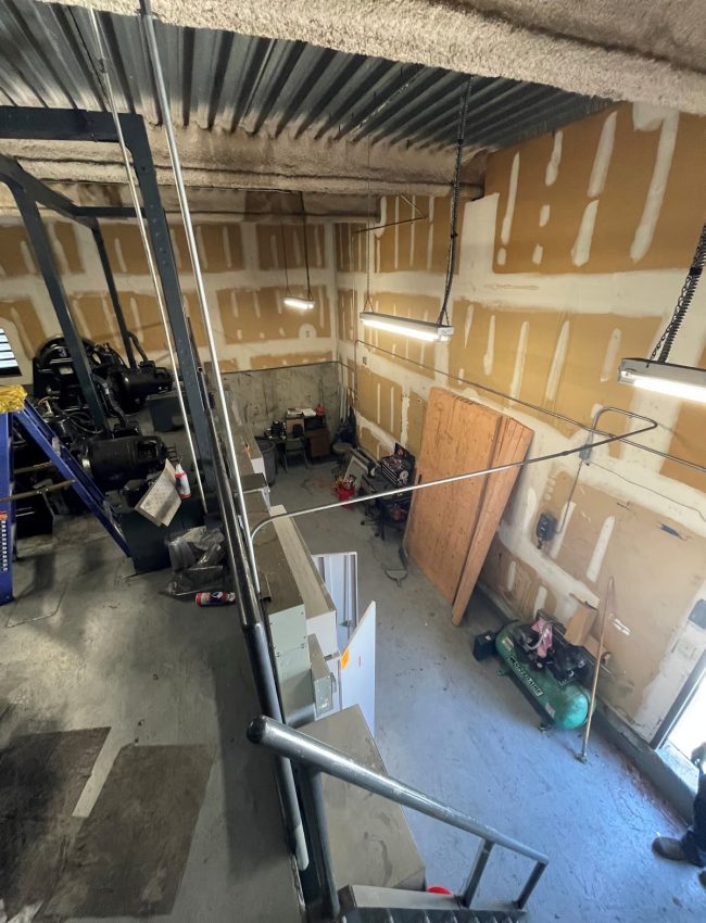 Elevator machine room with machinery, exposed wall panels, and overhead conduit.