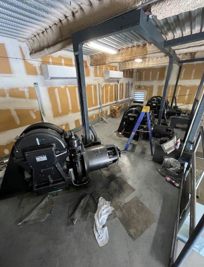 Elevator machinery beside exposed wall panels and overhead conduit.