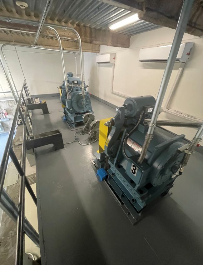 Two elevator machines in a room with finished white walls and a gray floor.